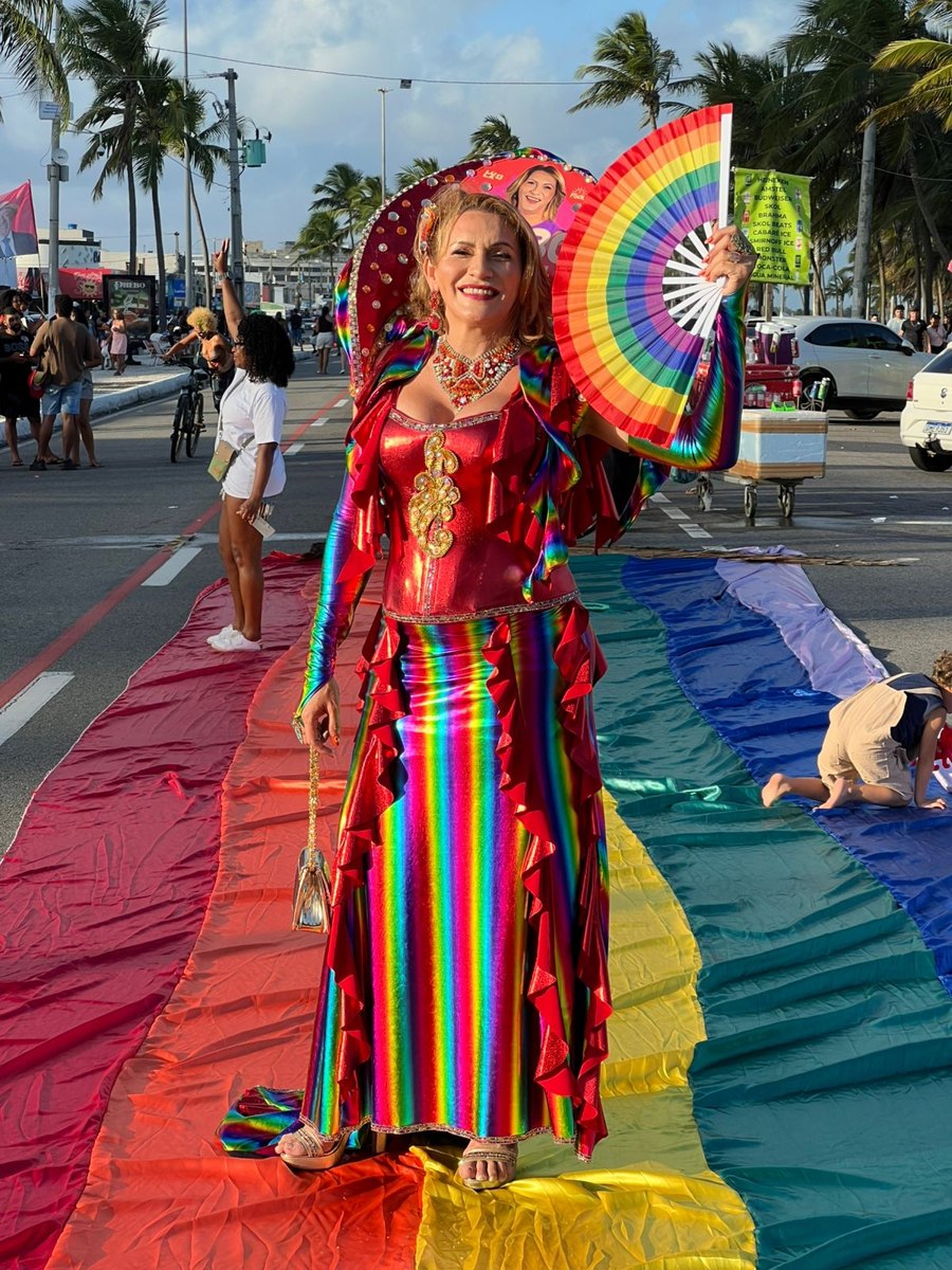 Linda Brasil com traje colorido sobre a bandeira do arco-íris na Orla de Atalaia